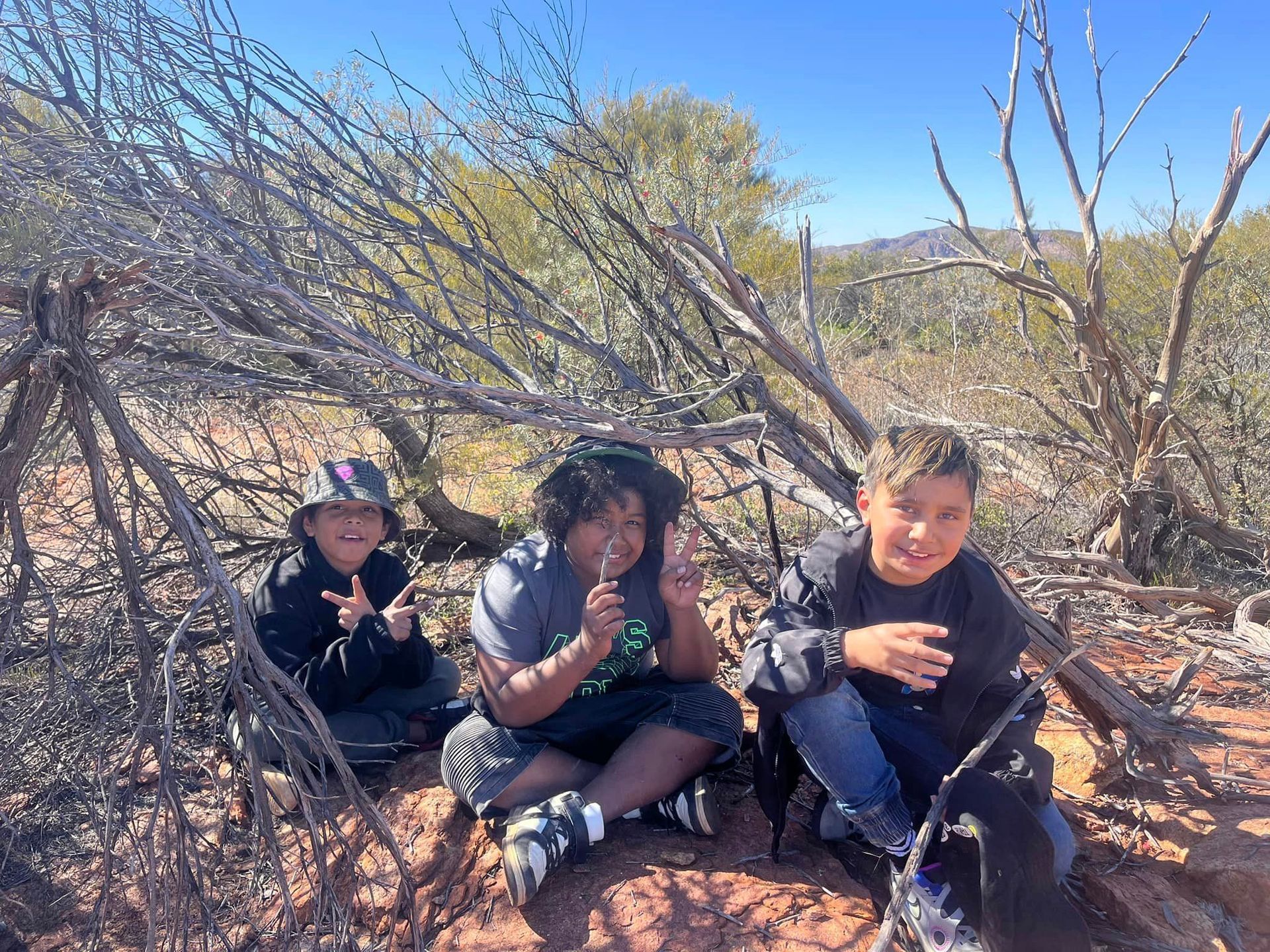 Three children sit under a bush in a desert landscape; they smile, and one licks a lollipop.