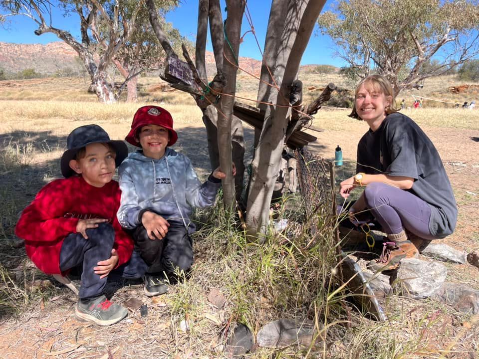 Three children and a woman crouch by a wooden structure in a field; mountains in the background.