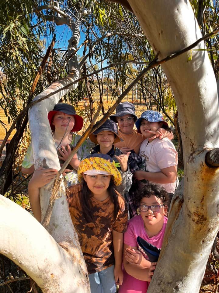 Group of children in hats, posing together behind a tree in an outdoor setting.