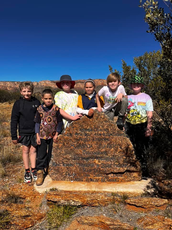 Six children pose around a large, brown rock outdoors on a sunny day.