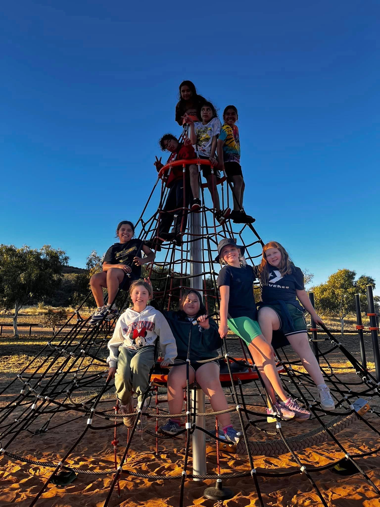 Children climbing a rope playground structure under a bright blue sky.