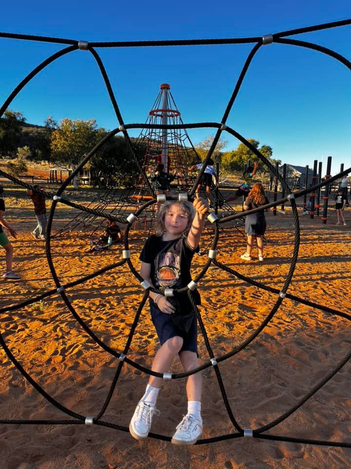 Boy smiles, poses inside black rope climbing structure at playground. Other kids play in the background.
