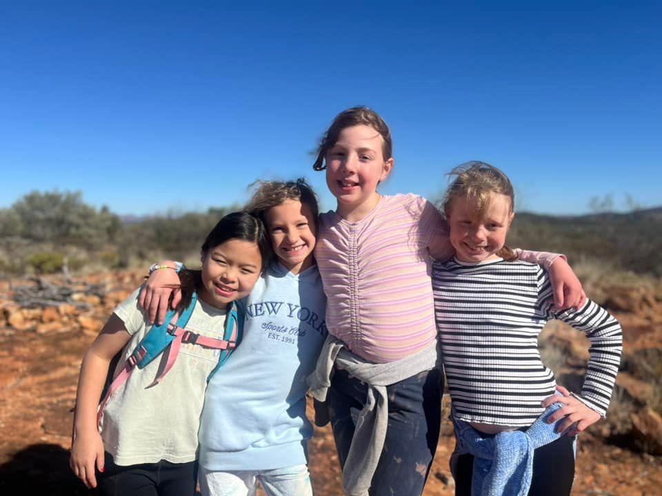Four smiling girls with arms around each other, outdoors on a sunny day.