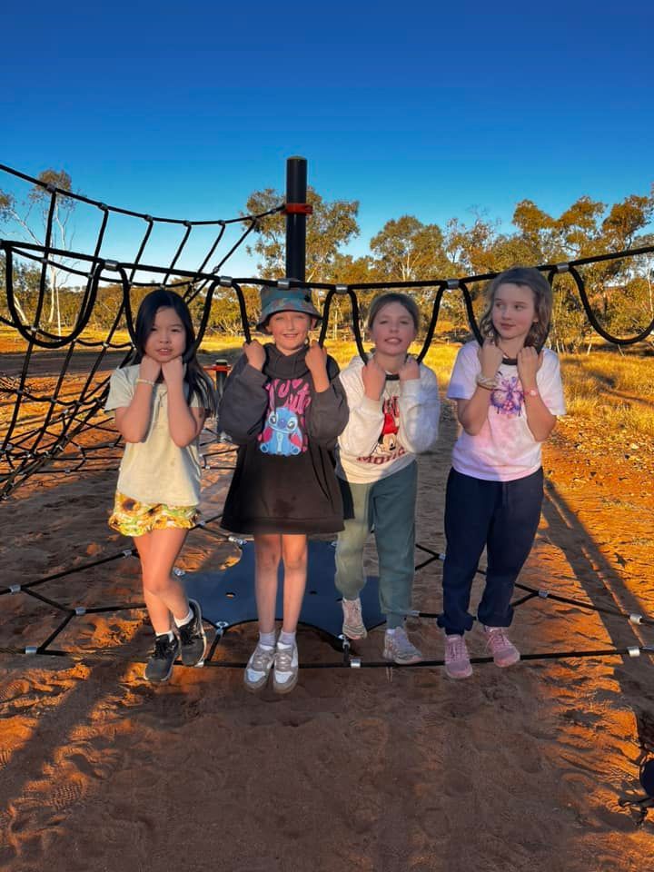 Four children smiling, posing near a climbing net in a park, blue sky overhead.