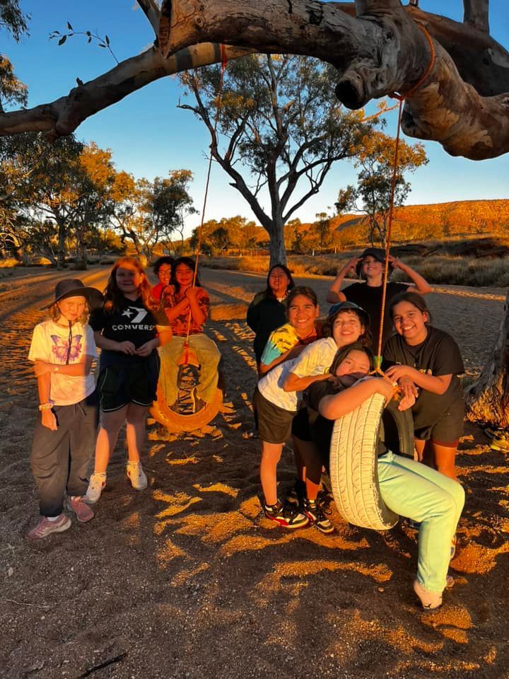 Group of teens smile by a tire swing suspended from a tree in a sunny outdoor setting.