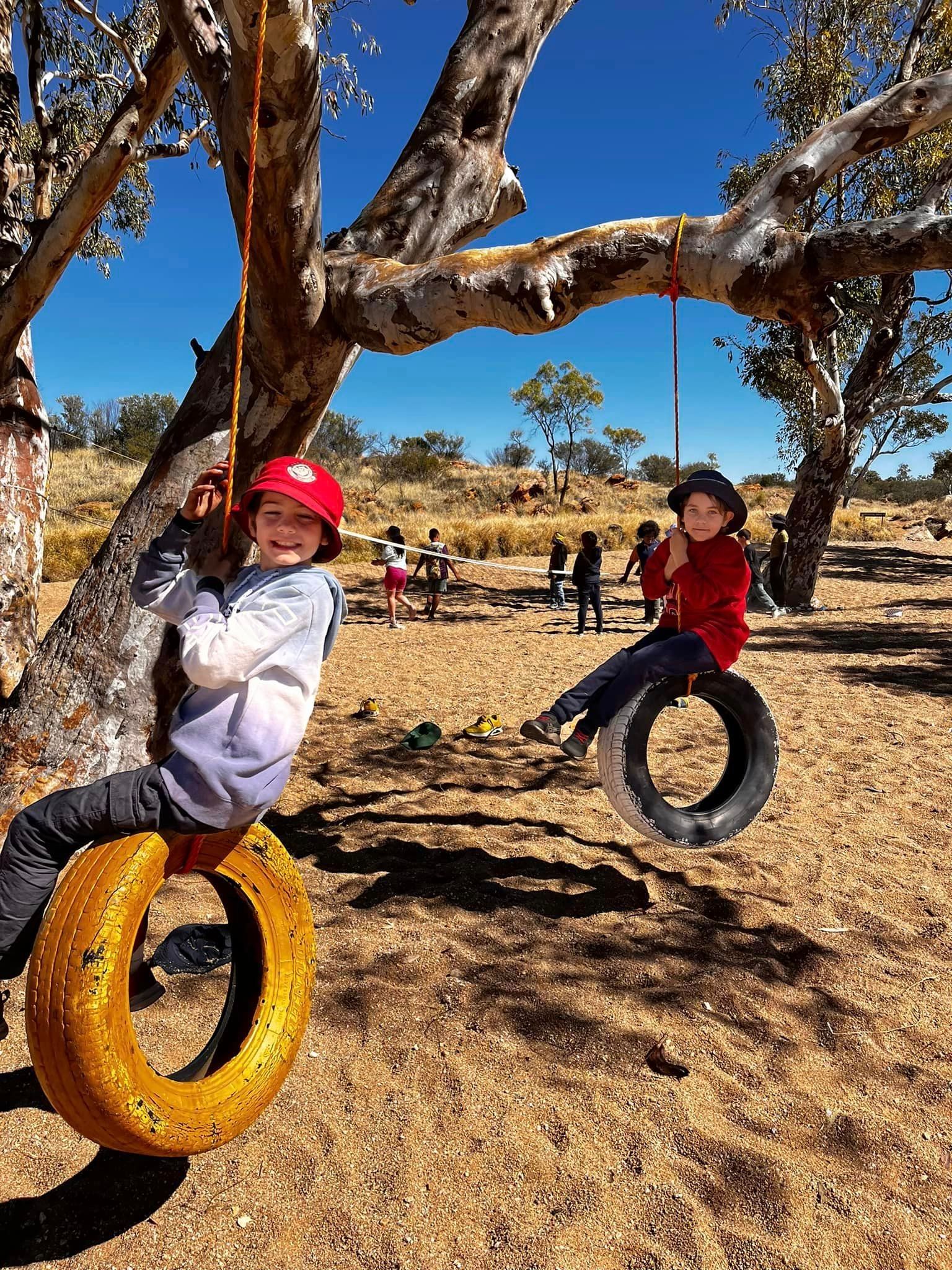 Two children on tire swings beneath a tree in a sunny field.