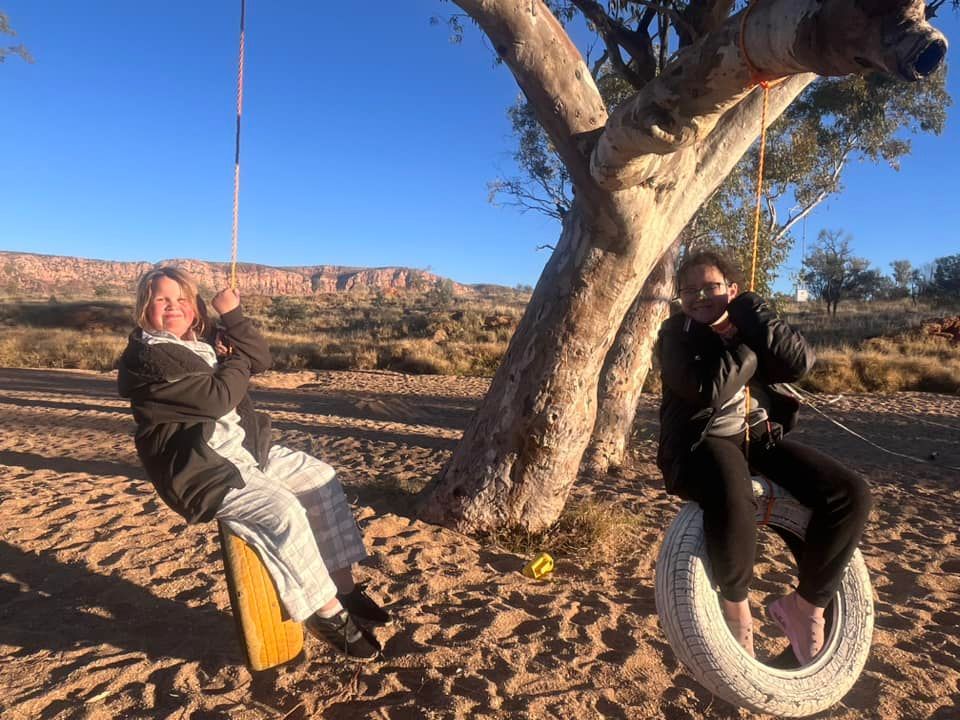 Two people swinging on tire swings in desert, smiles, blue sky, trees, distant red cliffs.