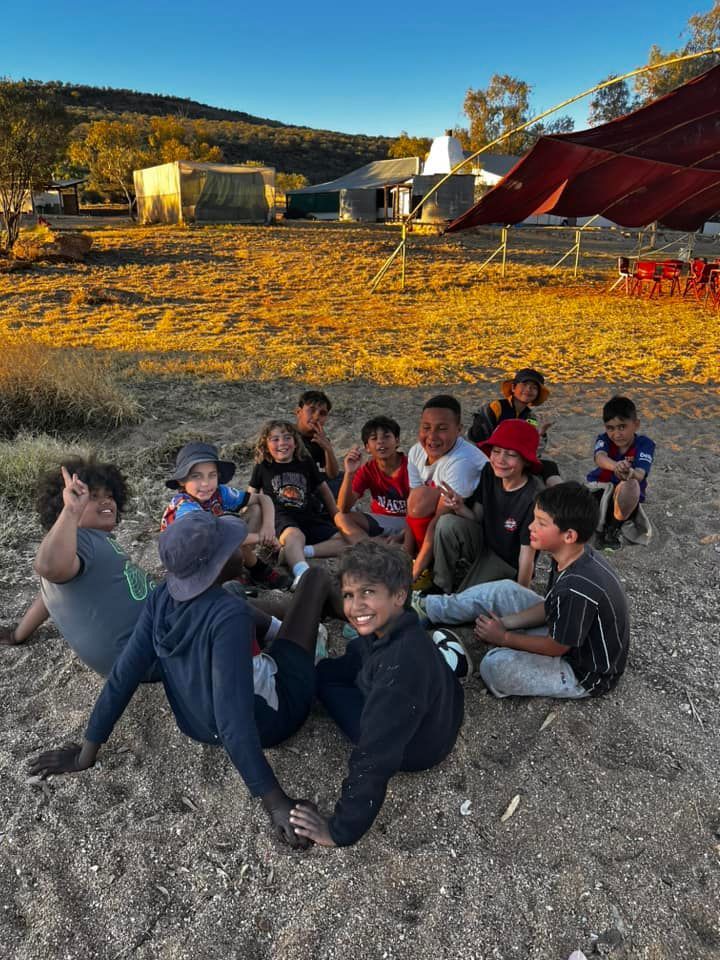 A group of children sits on a sandy area near a campsite; they smile and laugh, basking in warm sunlight.