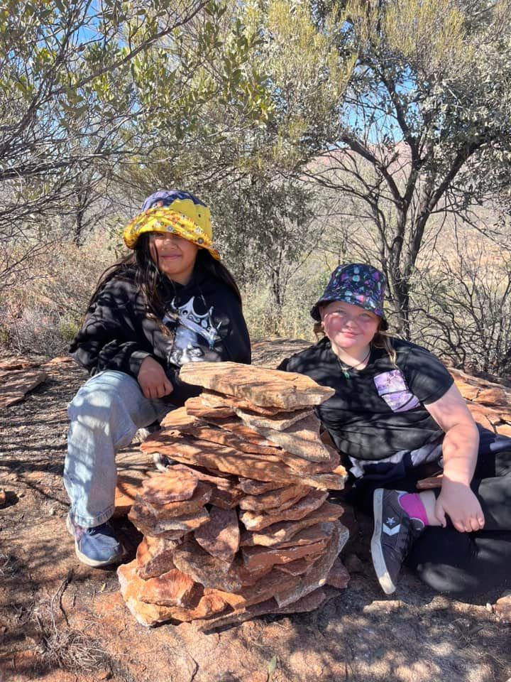 Two kids with hats sit near a rock cairn outdoors in the sun.