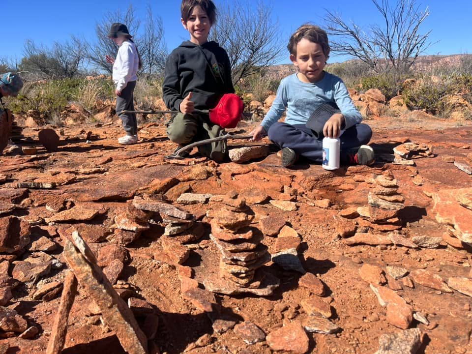 Children build stone cairns on red rock. One smiles, holding a stick with a red object; another looks on.