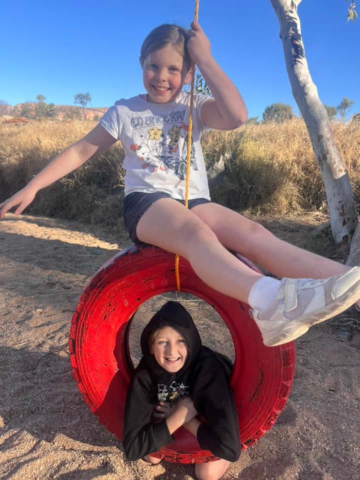 Two children playing on a red tire swing outdoors under a blue sky.