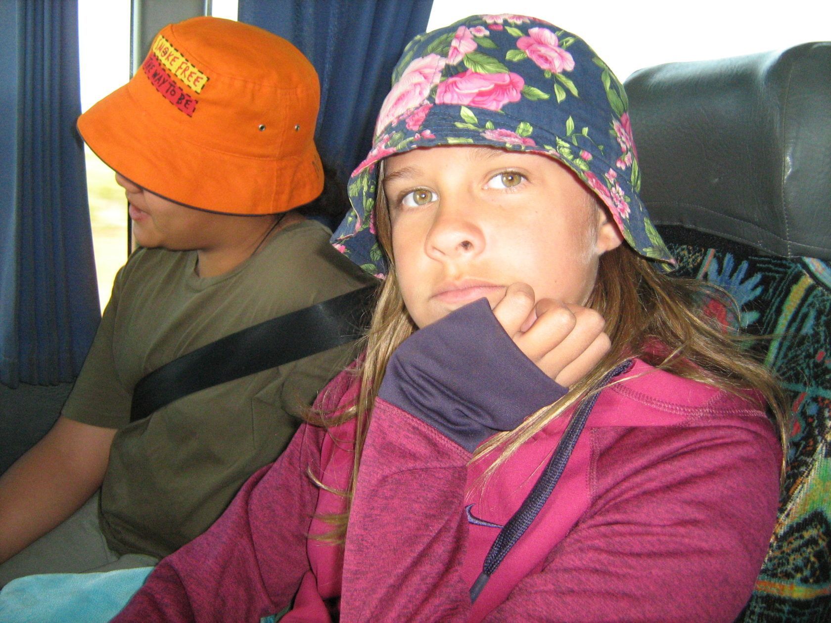 Girl in floral hat, resting chin on hand, beside person in orange hat, on a bus.