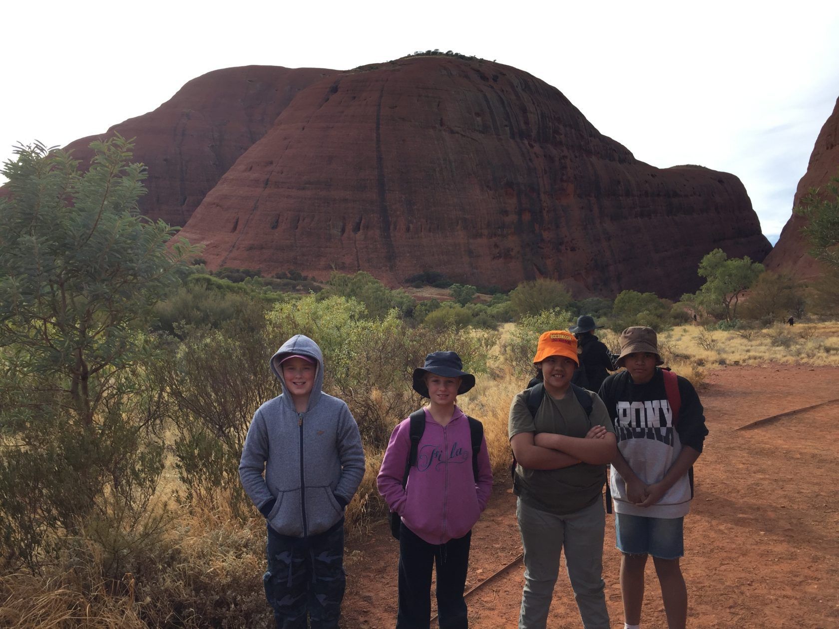 Four children stand in front of a large red rock formation in a desert landscape.