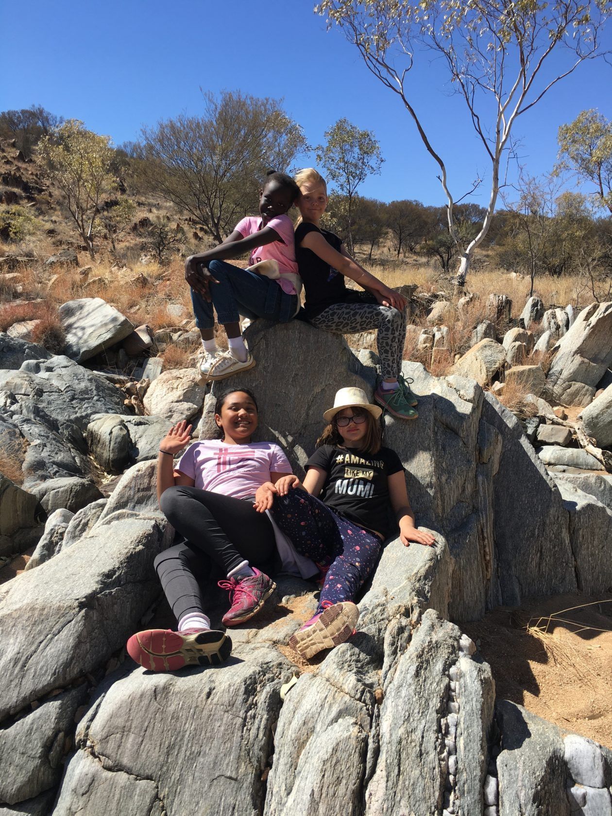 Four girls posing on rocks in a sunny, arid landscape. Two are seated high, two are seated below.