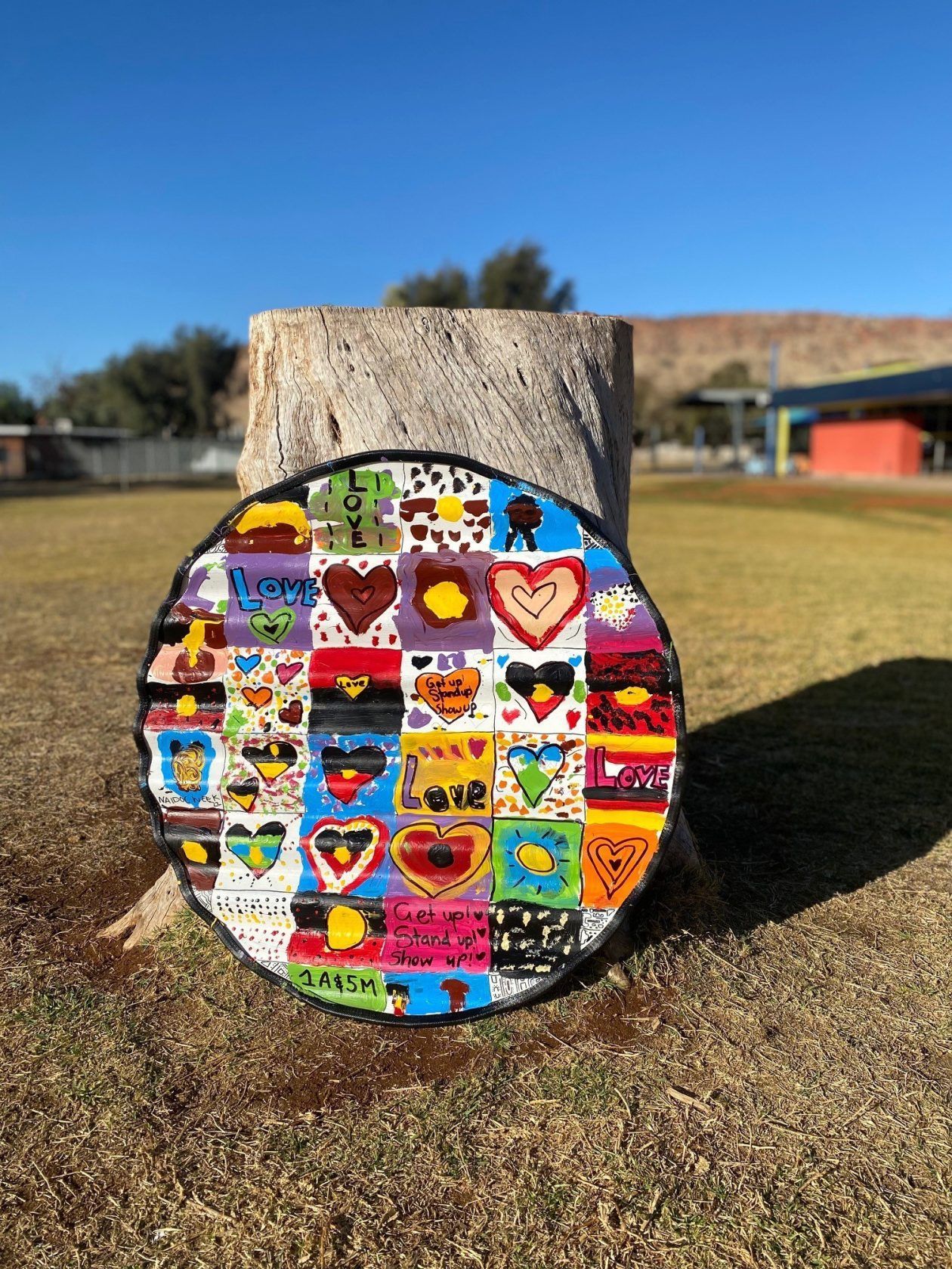 Circular artwork on a tree stump with colorful heart and Aboriginal flag designs in an outdoor setting.