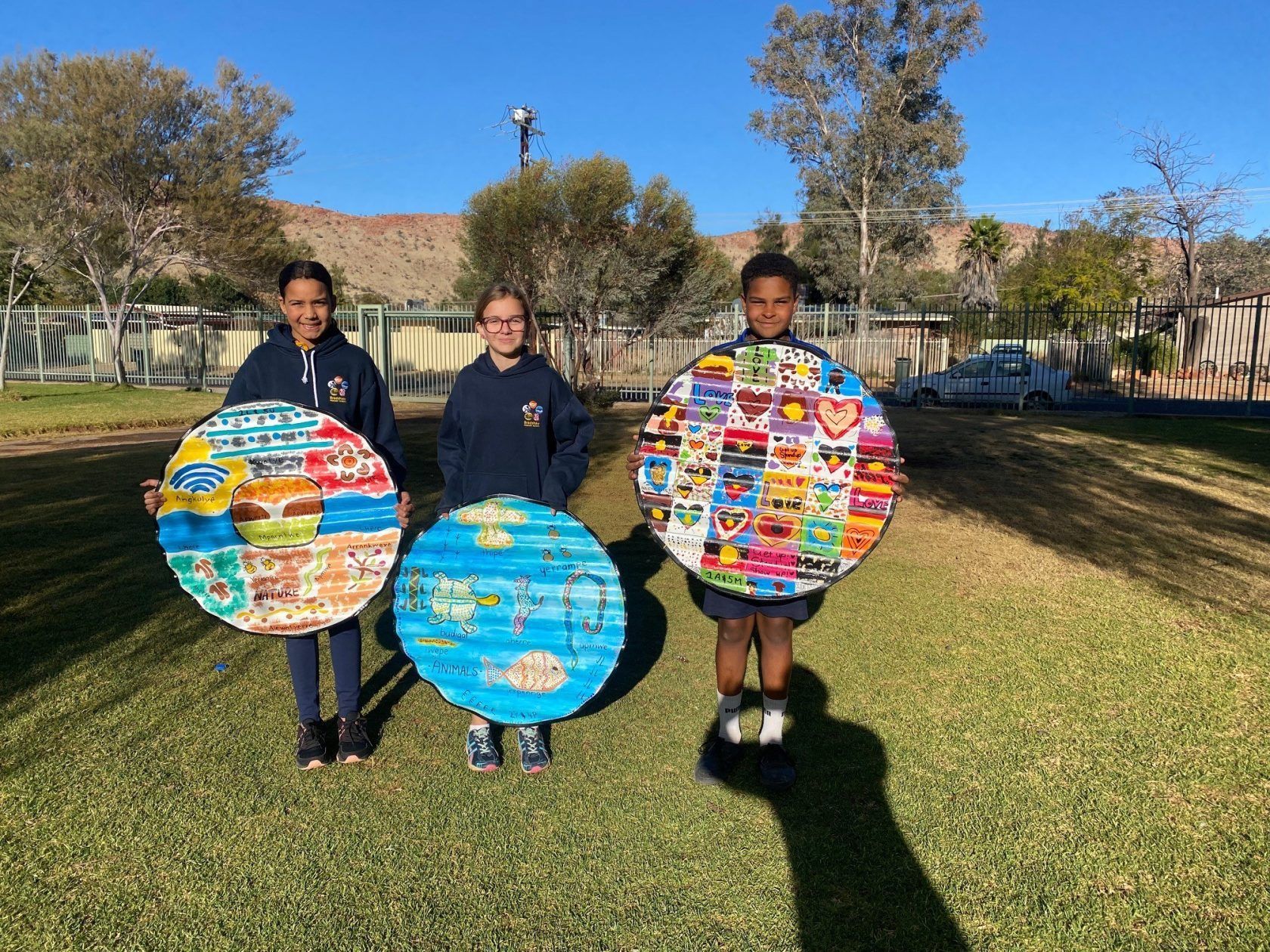 Three students holding colorful, circular artwork on a grassy field, with a fence and buildings in the background.