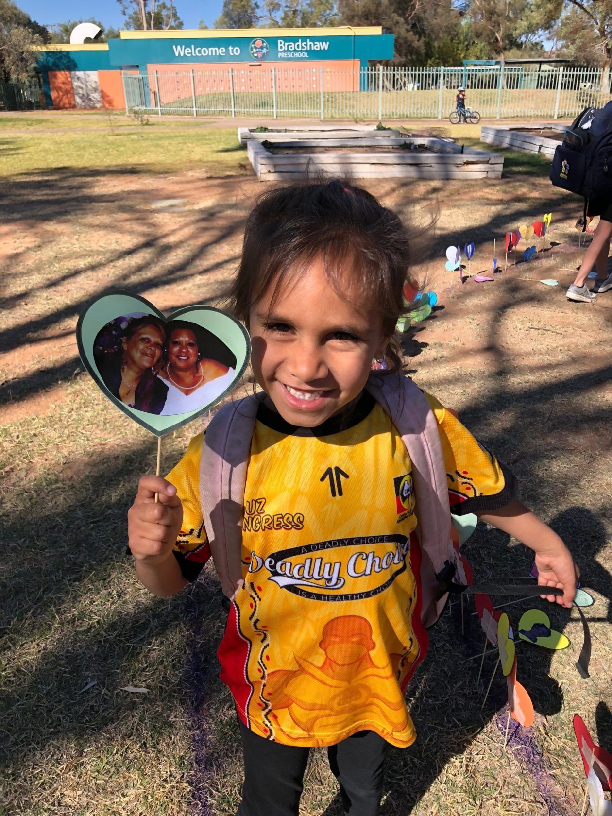 Girl smiling, holding a heart-shaped photo, wearing a yellow jersey, outside a school building.