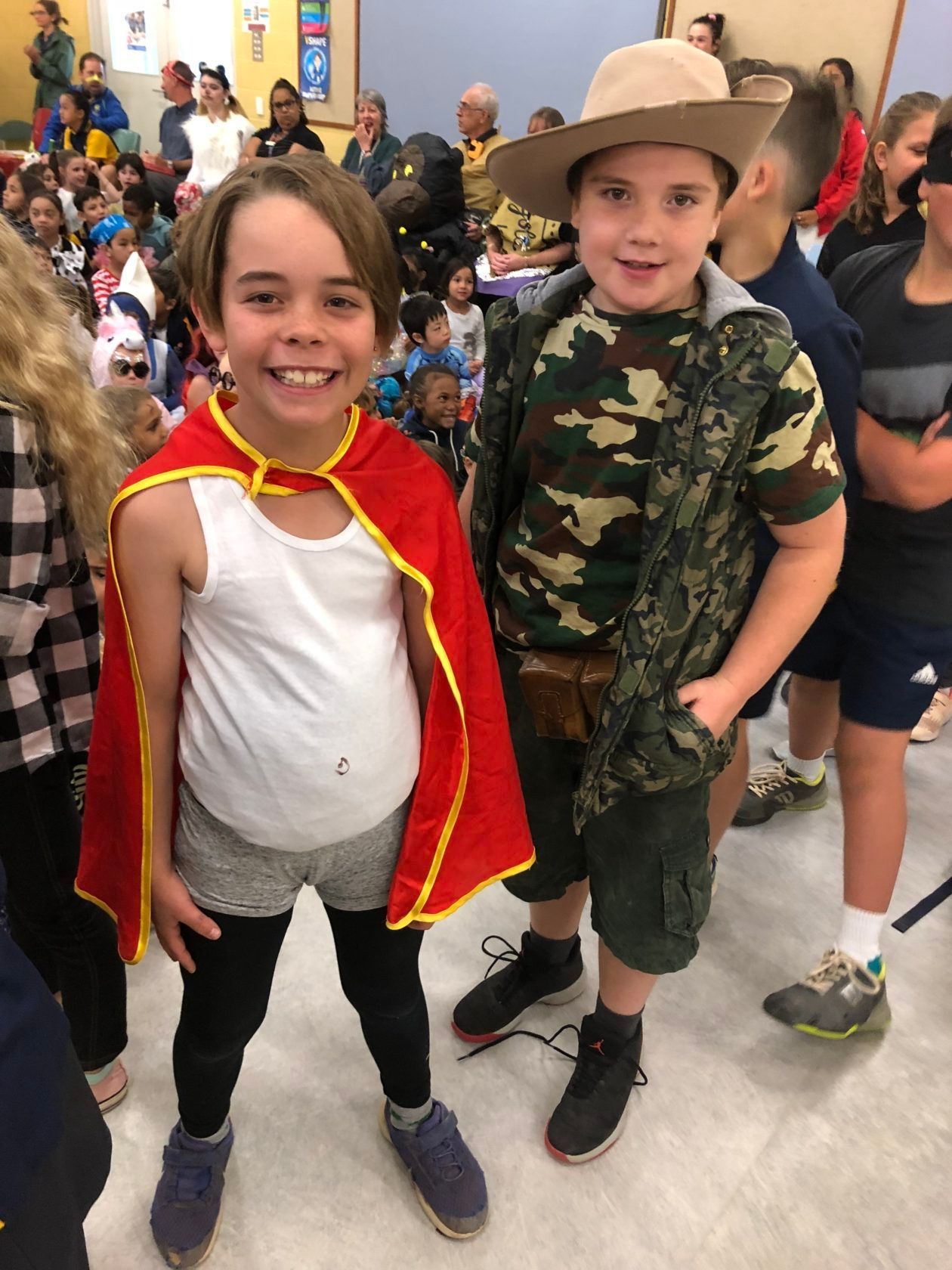 Two children in costumes. One wears a cape and the other a safari outfit in a room with a crowd.