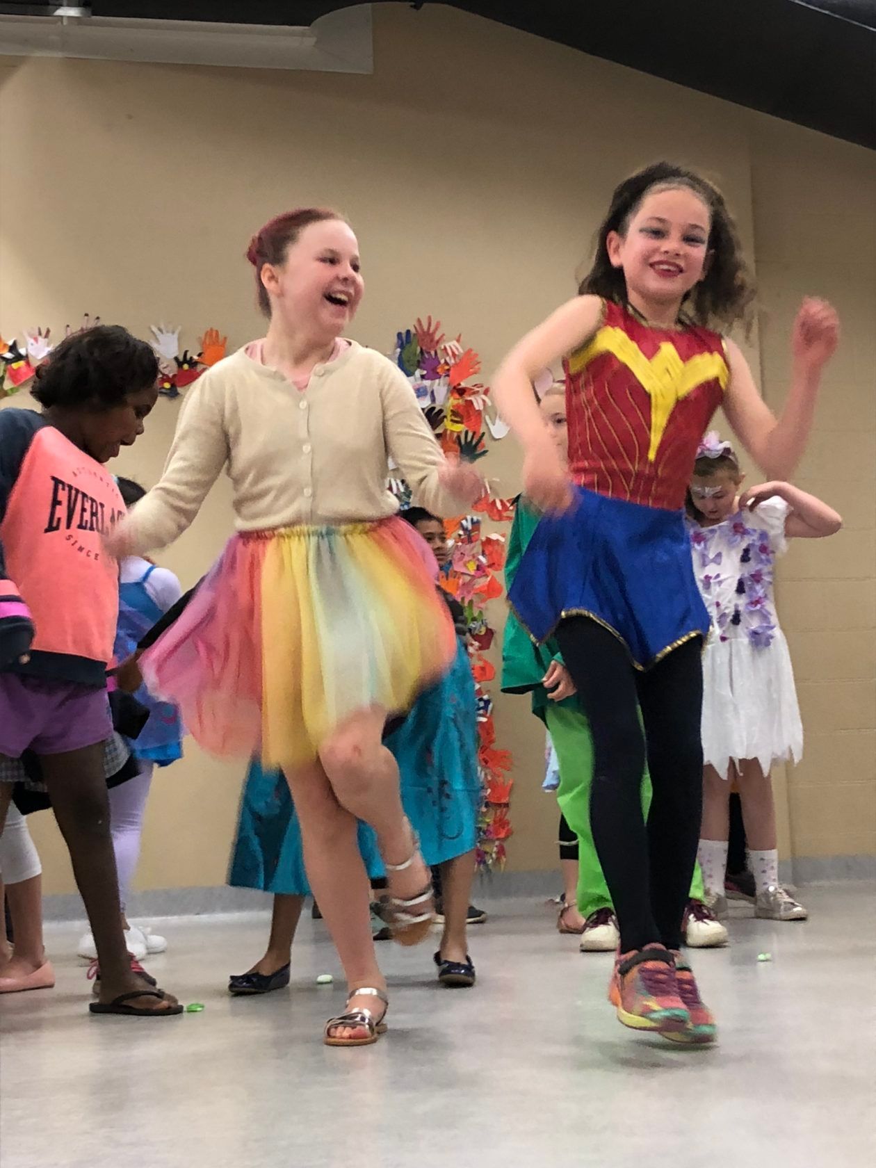 Children dancing in a room. A girl in a Wonder Woman costume dances next to a girl in a rainbow skirt.