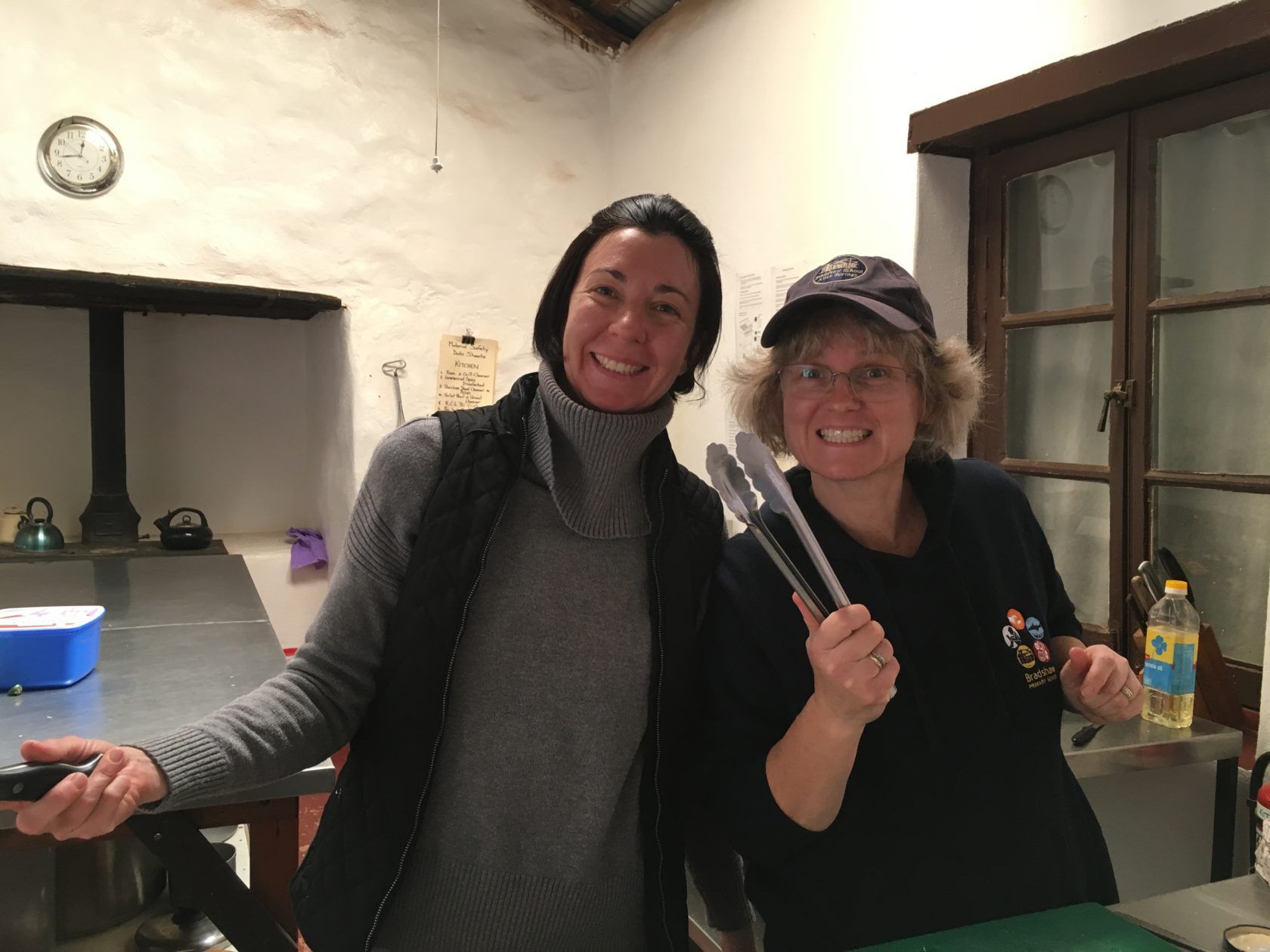 Two smiling women in a kitchen. One holds tongs, the other a knife.