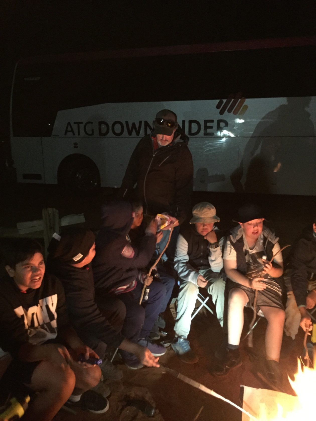 A group of children sitting around a campfire at night, with an adult standing nearby in front of a bus.