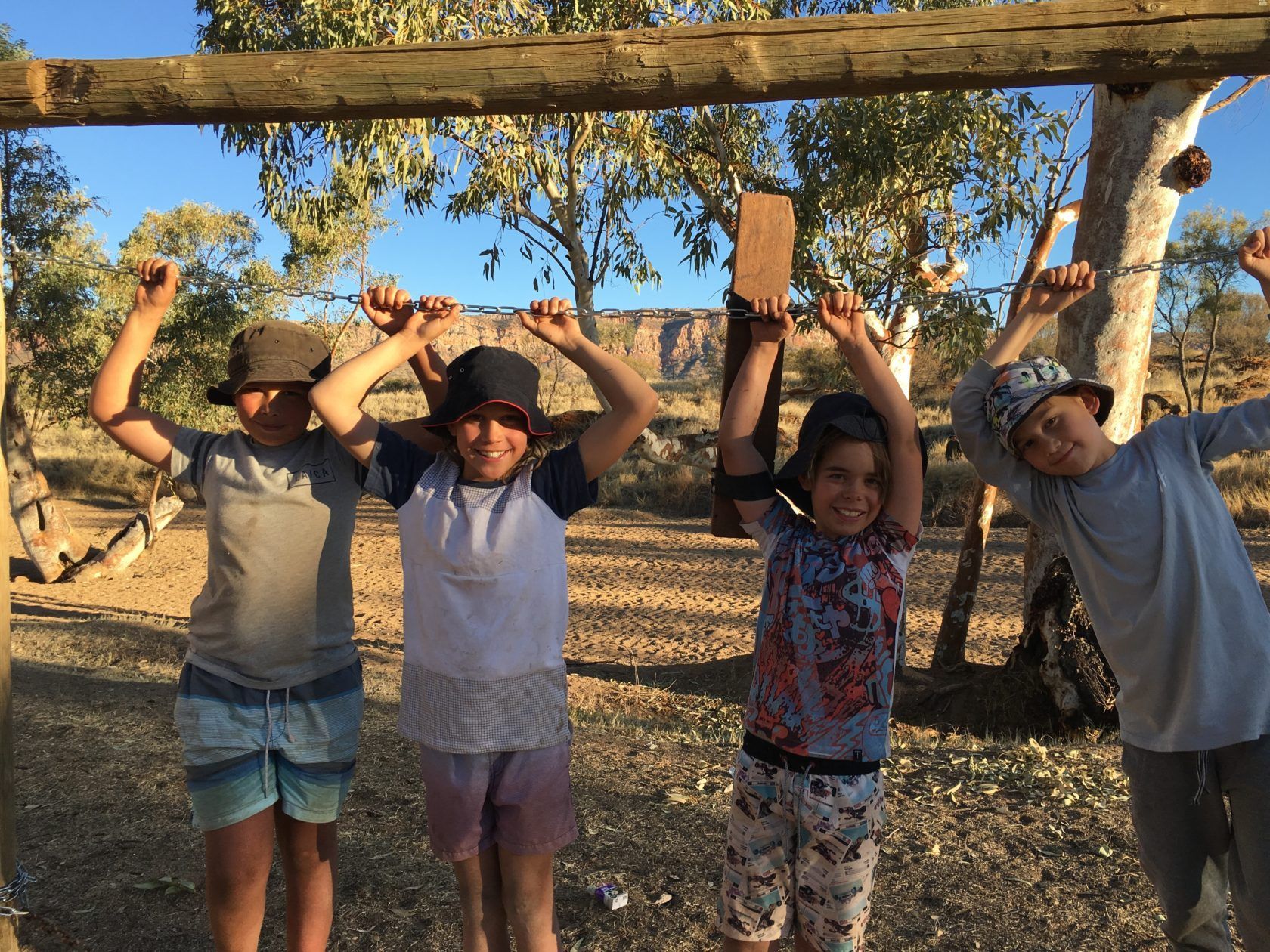 Four children in hats raise their arms, holding barbed wire under a wooden beam in a desert setting.