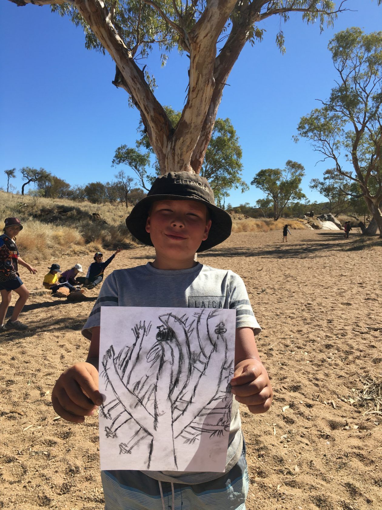 Boy in hat holds drawing of tree; outdoors in sunny, dry landscape. Others in background.