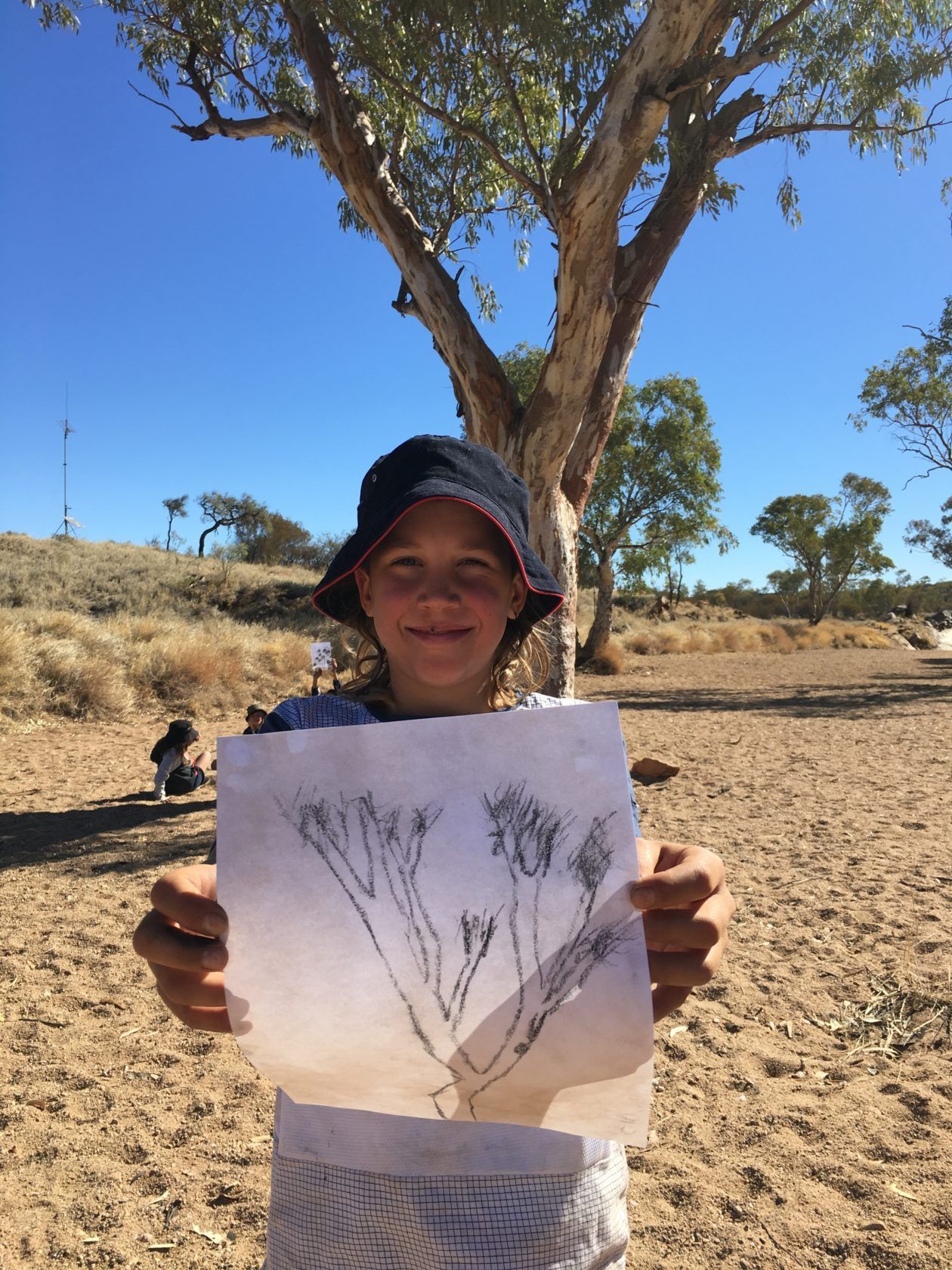 Girl in hat holding drawing of tree in sunny desert setting.