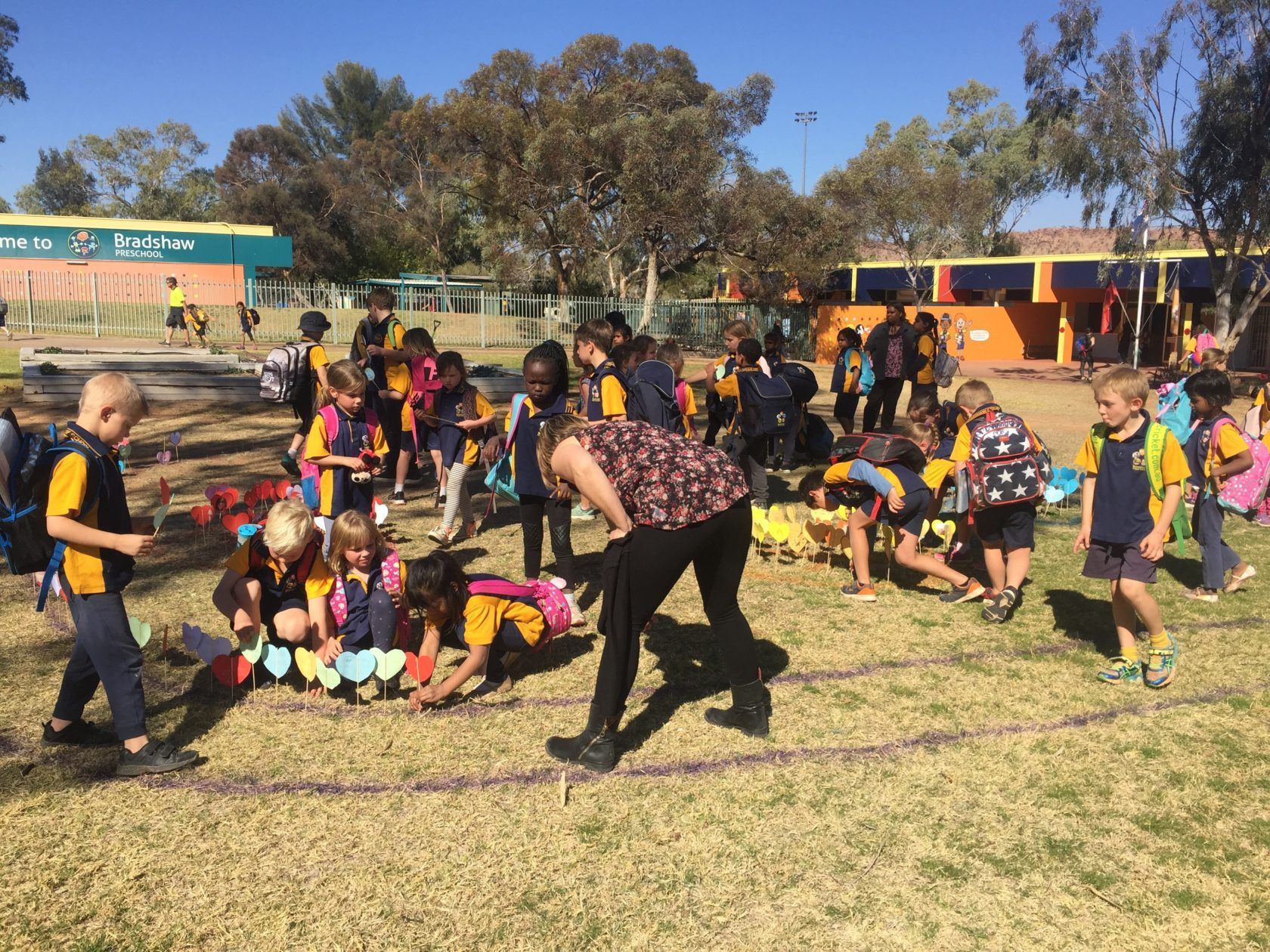 Children and teacher on a grass field, circling with string and pencils. Trees and school buildings in background.