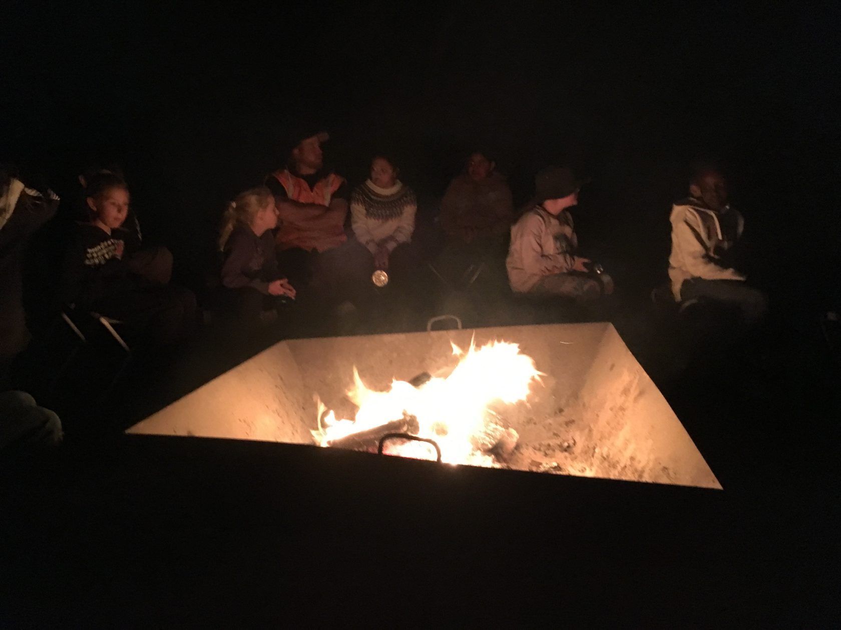 People sitting around a fire pit at night. Flames glow in the square metal pit, with dark figures watching.