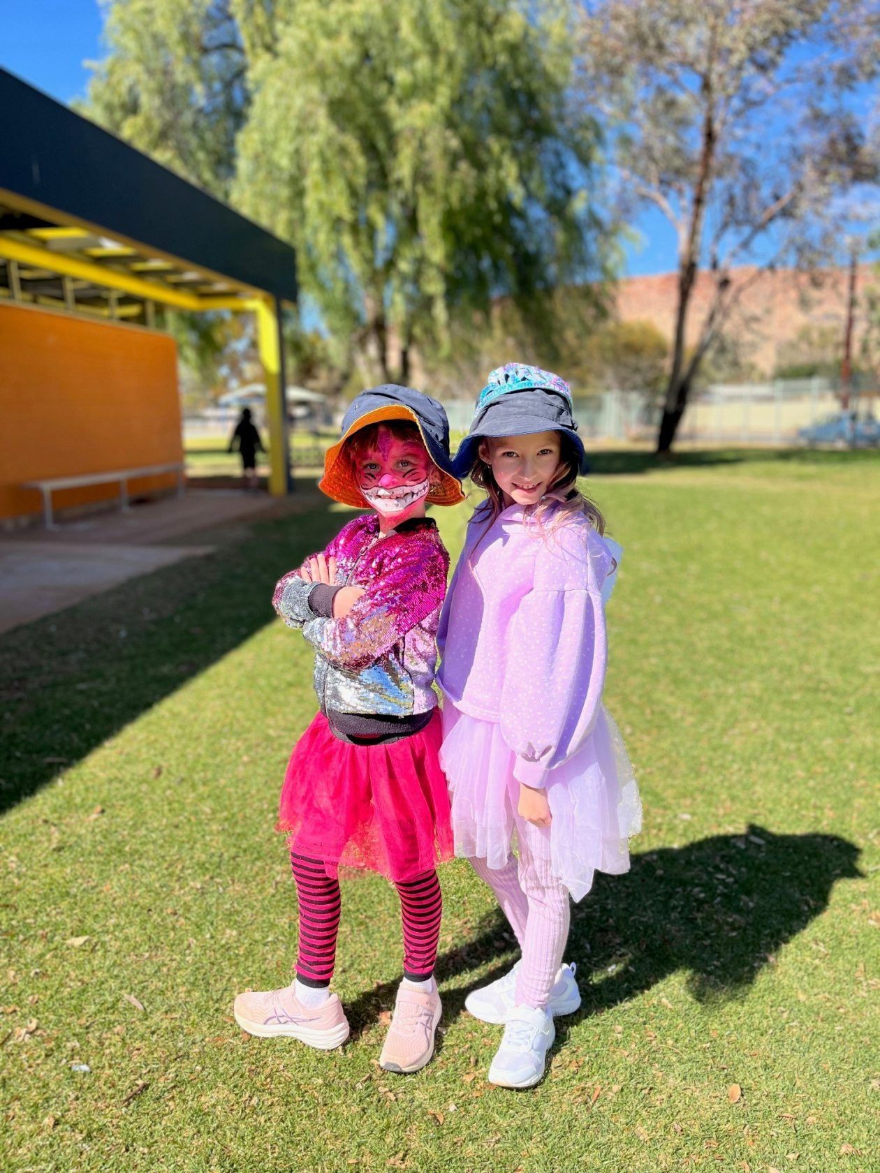 Two girls in costume pose on green grass, one with face paint and arms crossed, the other in a light purple outfit.
