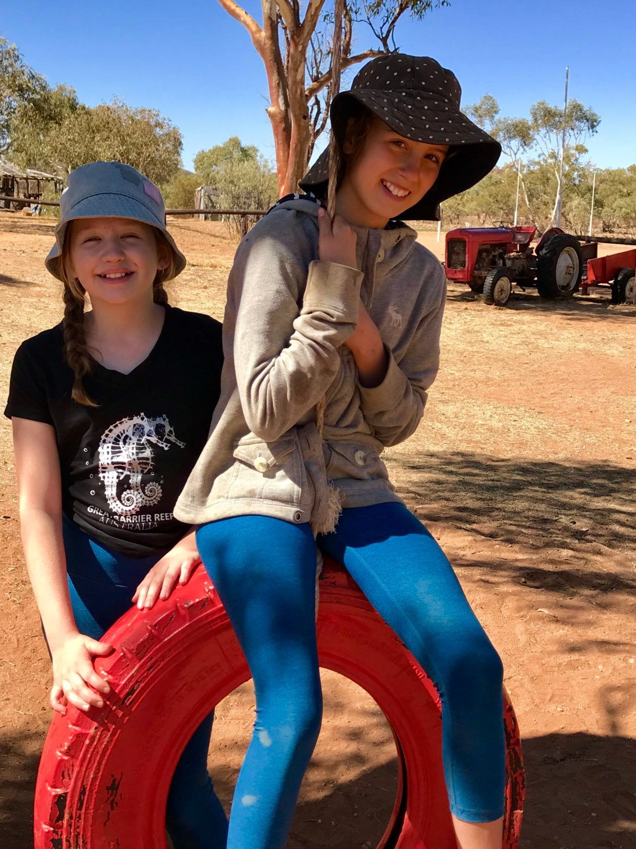 Two girls wearing hats, one on a red tire. They are smiling in a sunny outdoor setting.