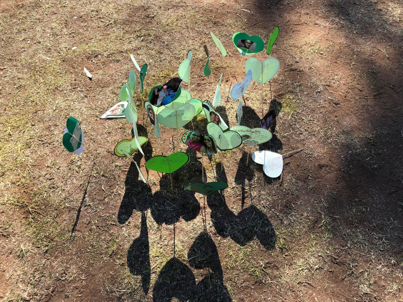 Overhead shot of a whimsical art installation with green, blue, and white paper leaves and shadows on brown ground.