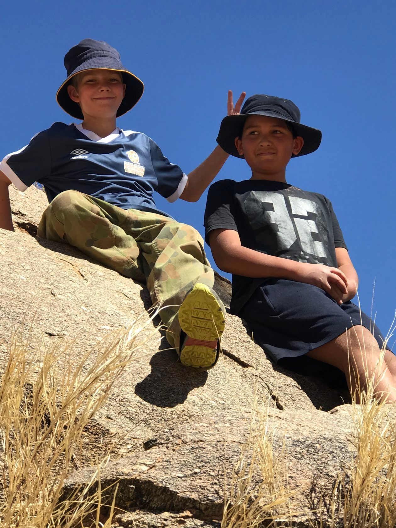 Two boys in hats sitting on a rock, one makes a peace sign. Blue sky backdrop.
