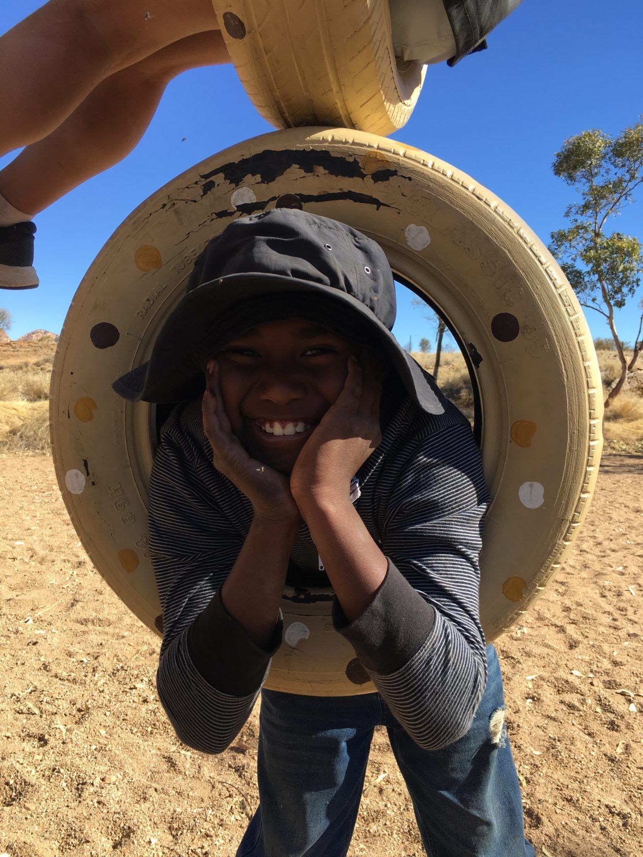 A smiling child wearing a hat, framed by a tire, with hands on their face. Outdoors, desert.