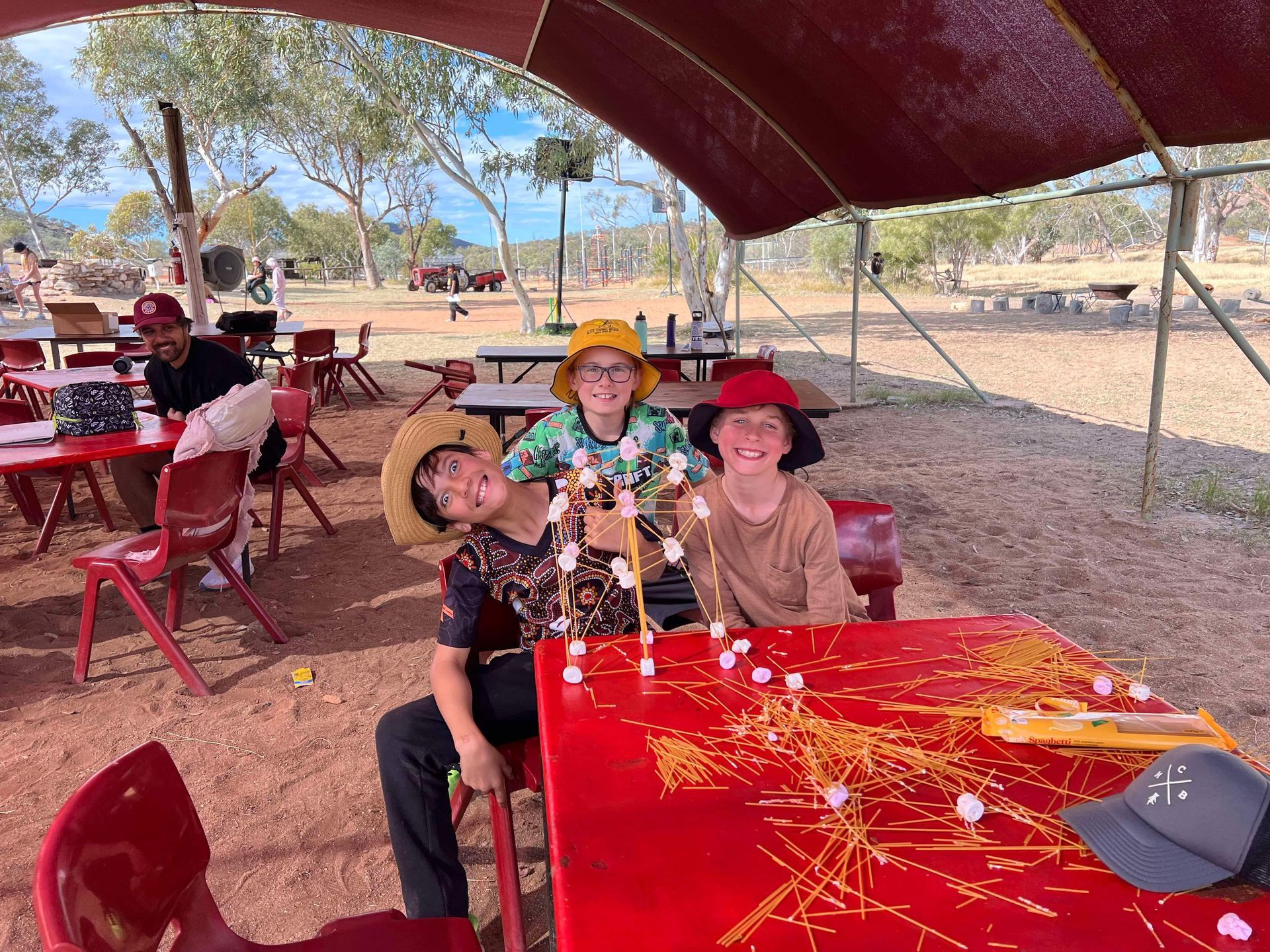 Kids building with spaghetti and marshmallows at a red table, under a shade.