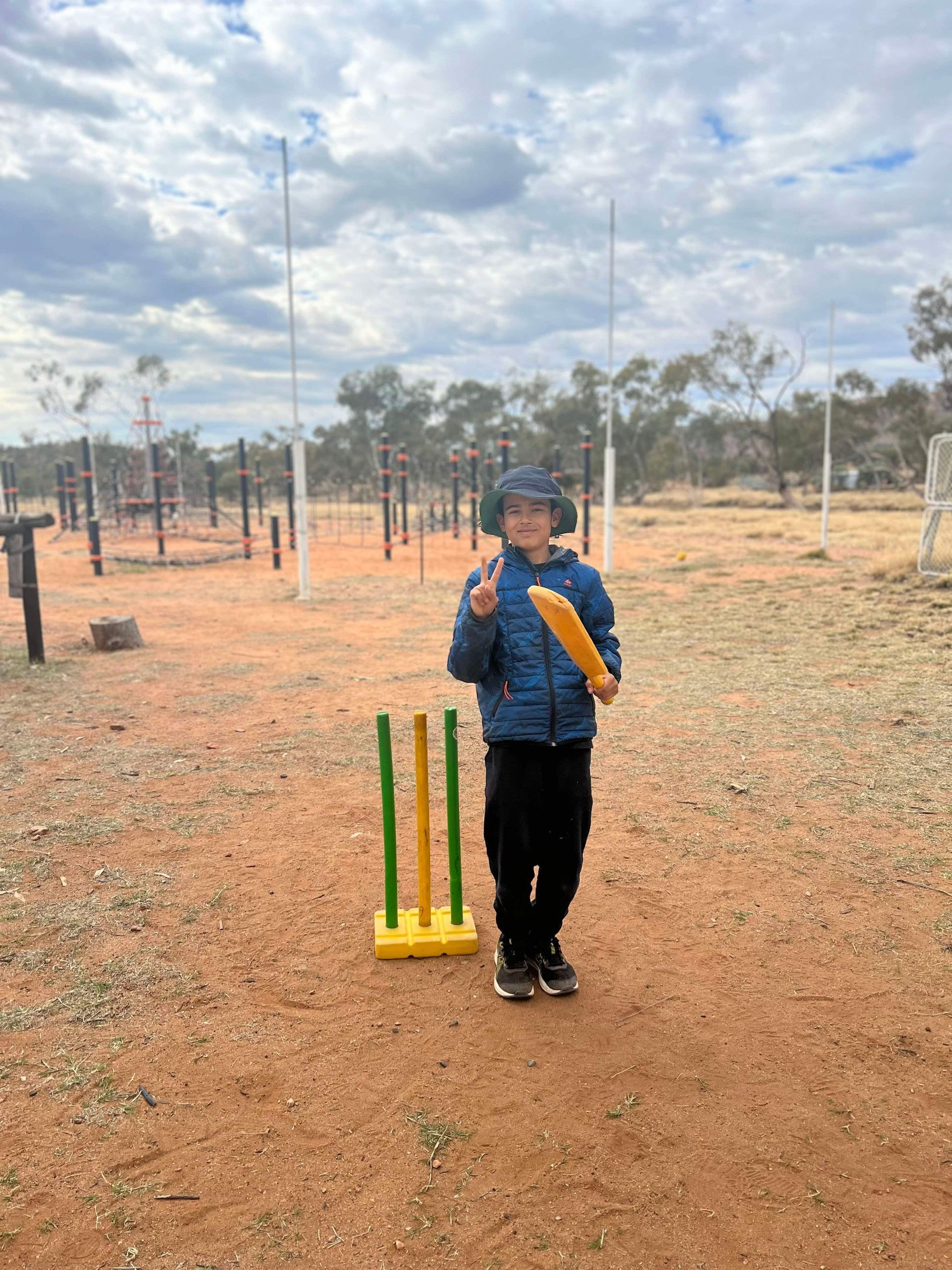Boy with cricket bat and wickets outdoors, smiling. Wearing blue jacket, black pants, on red dirt.
