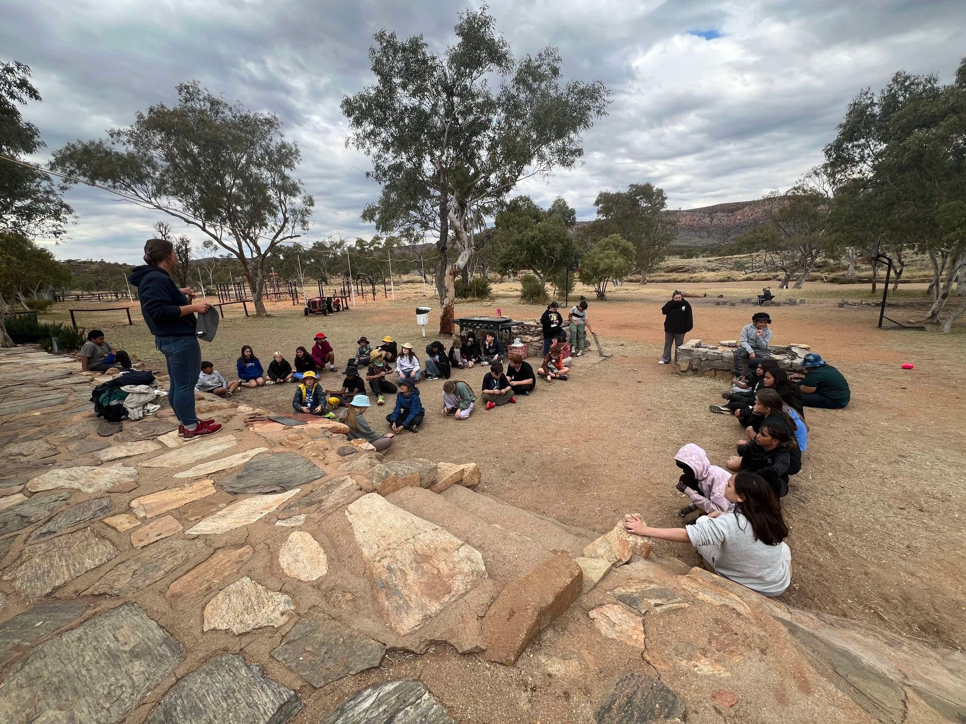 Group of people gathered outdoors, listening to a speaker on stone steps. Cloudy sky, red dirt, and trees in the background.