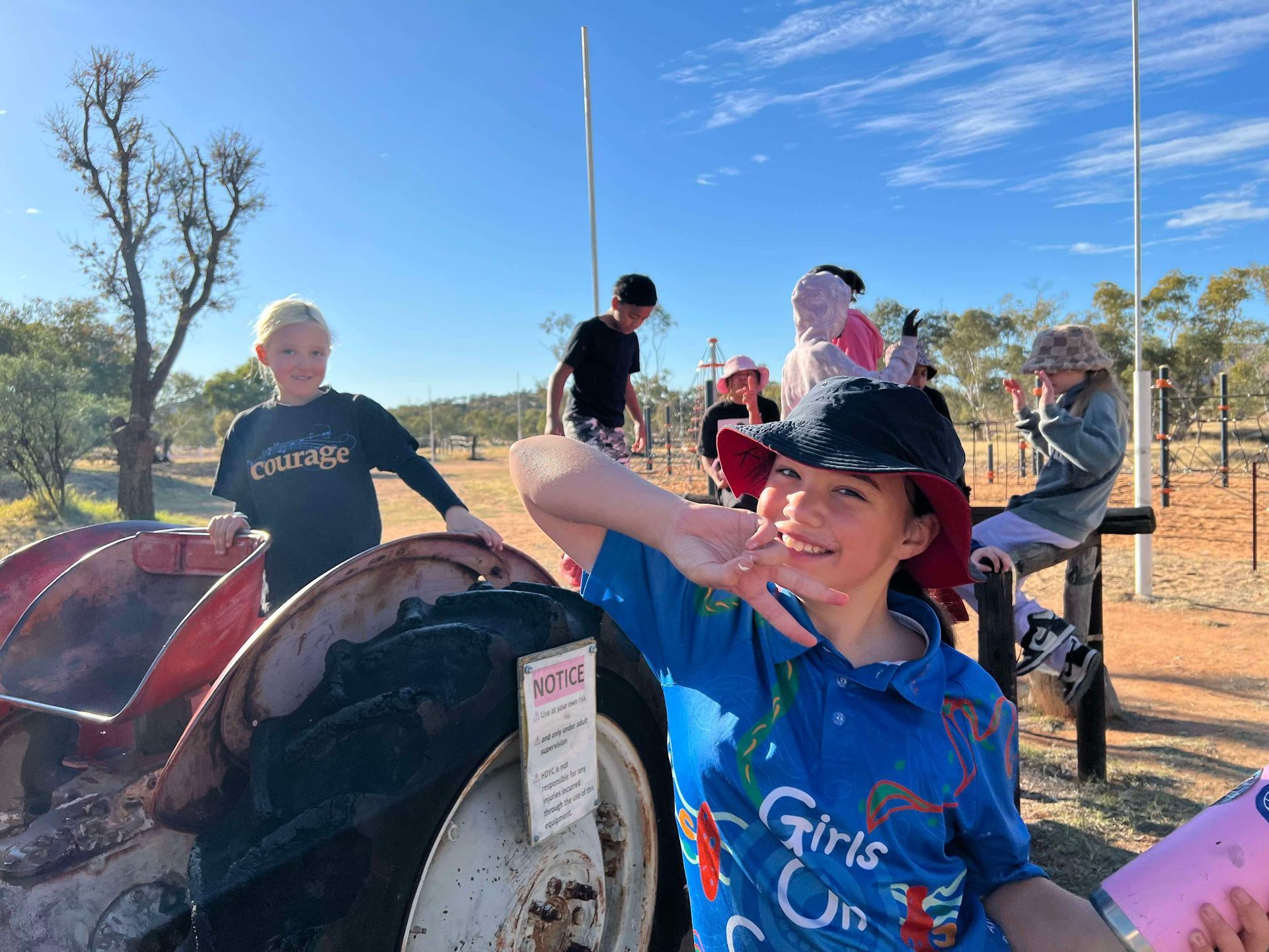 Children posing near an old tractor outdoors on a sunny day, some smiling, making peace signs.
