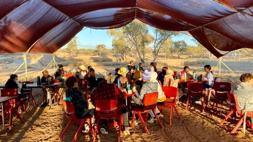 Children seated at red tables and chairs under a shaded tent outdoors; arid landscape in background.
