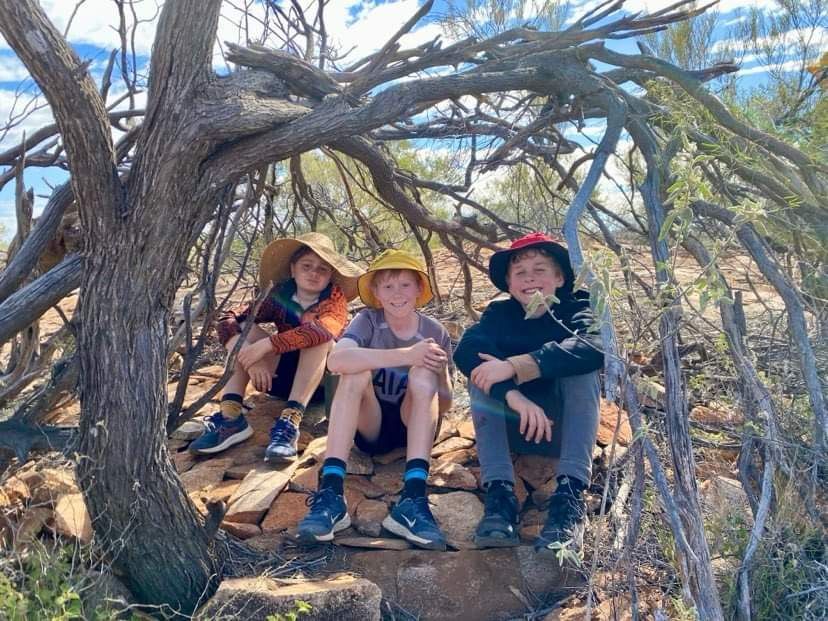 Three kids wearing hats sitting under a tree, smiling in a sunny outdoor setting.