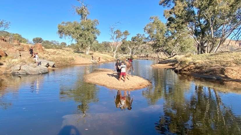 People stand on a small island in a river, trees surround the water in a sunny, outdoor setting.