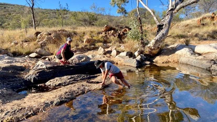 Two people exploring a shallow creek bed in a sunny, rocky landscape. One bends over, wading in water.