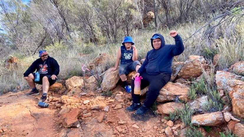 Three people rest on a rocky hillside. One raises a fist, another sits, and the last is relaxed.