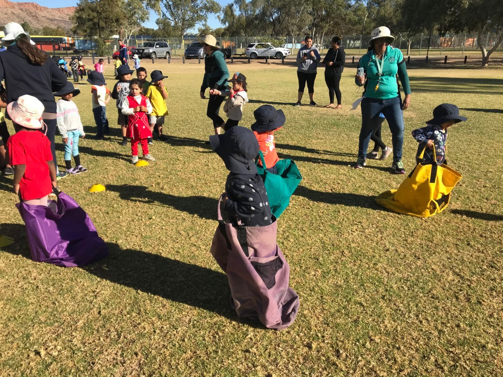 Children in colorful outfits playing a sack race game on a grassy field, overseen by adults.