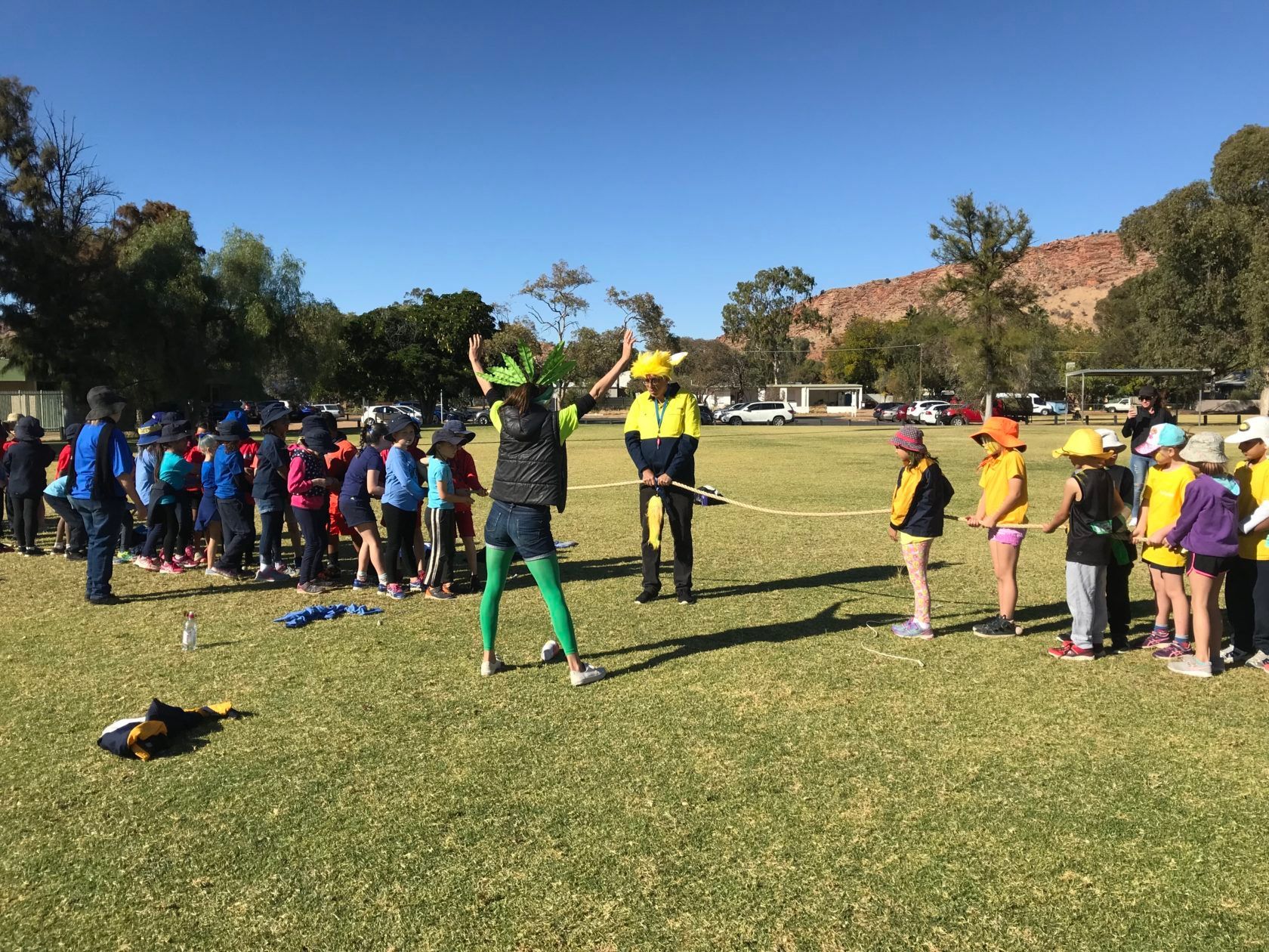 Children in colorful hats and a teacher do an outdoor activity on a green field.
