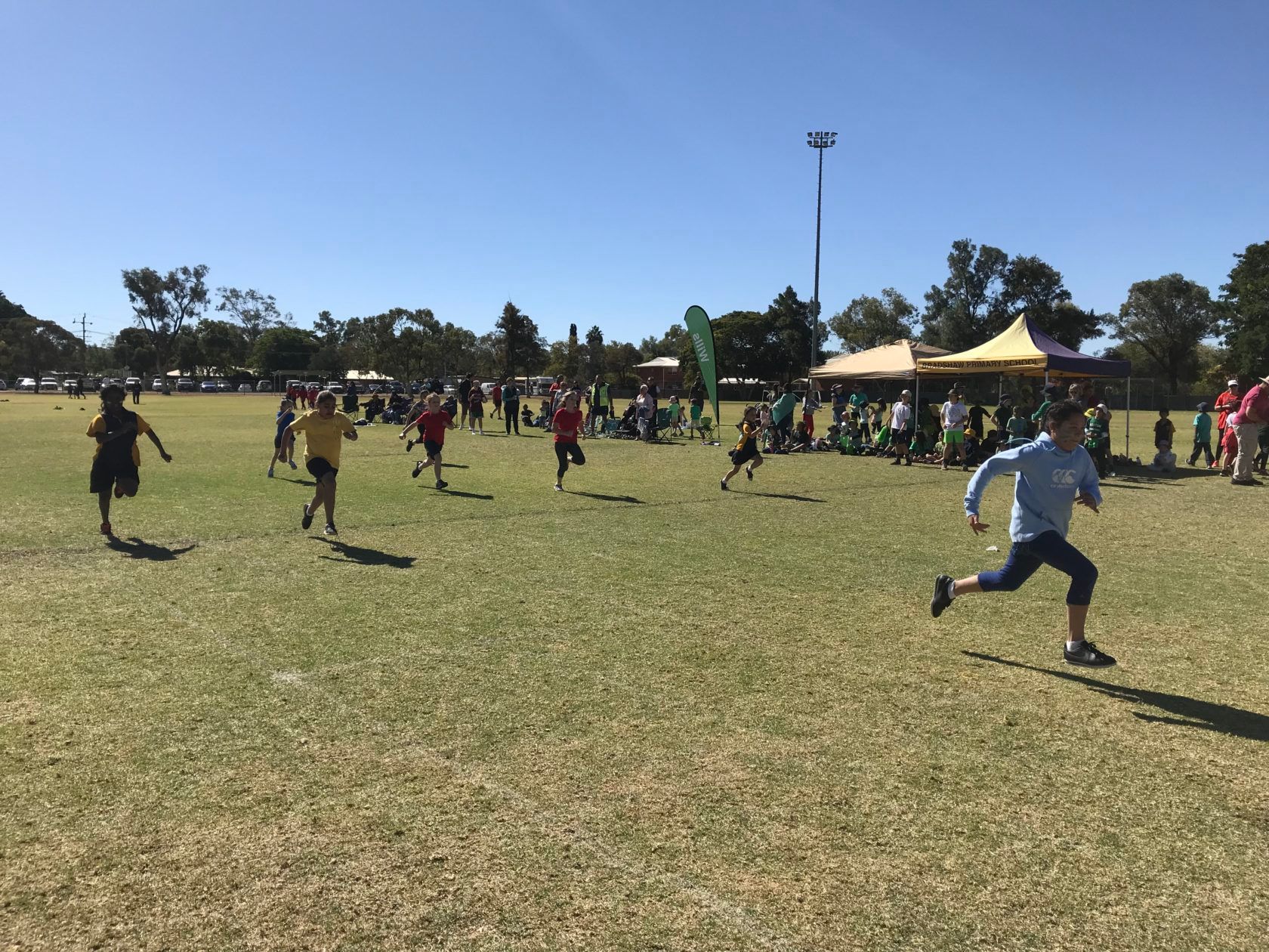 Children running a race on a grassy field under a clear blue sky. Spectators watch in the distance.
