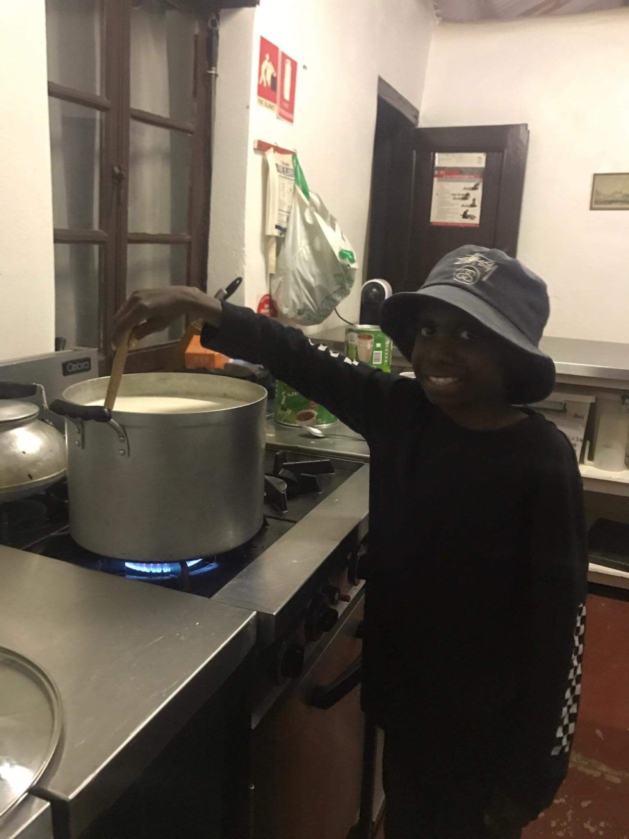 Young person stirring a pot on a stove in a kitchen, wearing a black outfit and a bucket hat.