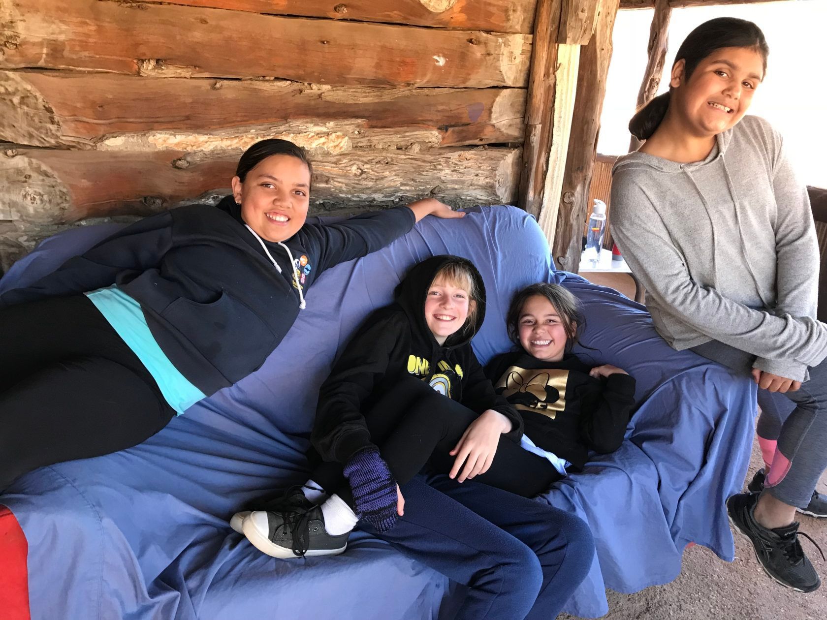 Four smiling children seated and leaning on a blue couch in a rustic, outdoor setting.