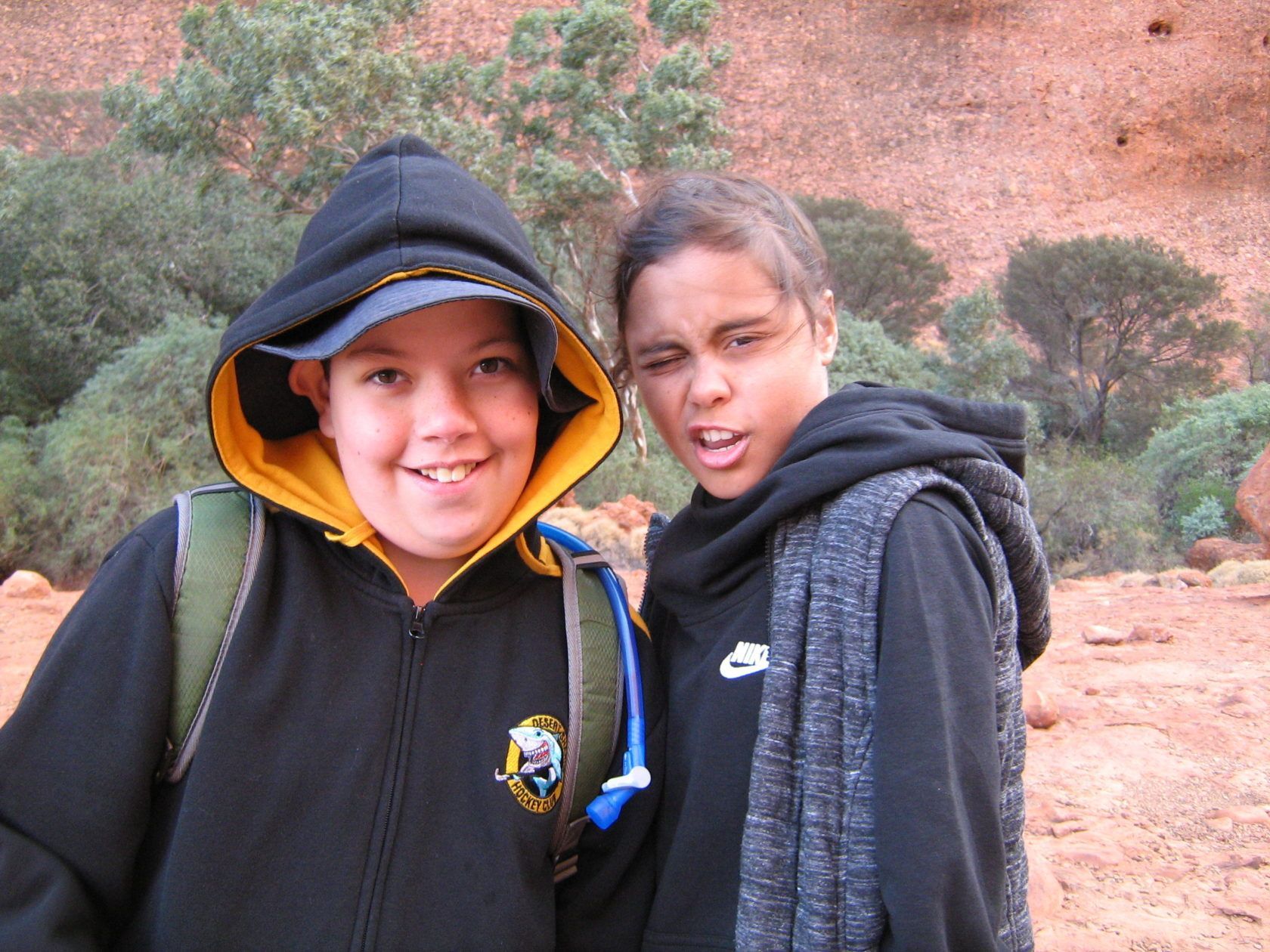 Two people in hoodies pose outdoors, smiling at the camera. Red rock and greenery in the background.